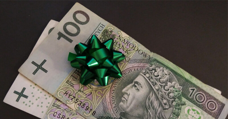 Dwa banknoty o nominale 100 złotych z błyszczącą zieloną kokardą na wierzchu, umieszczone na ciemnej powierzchni.
