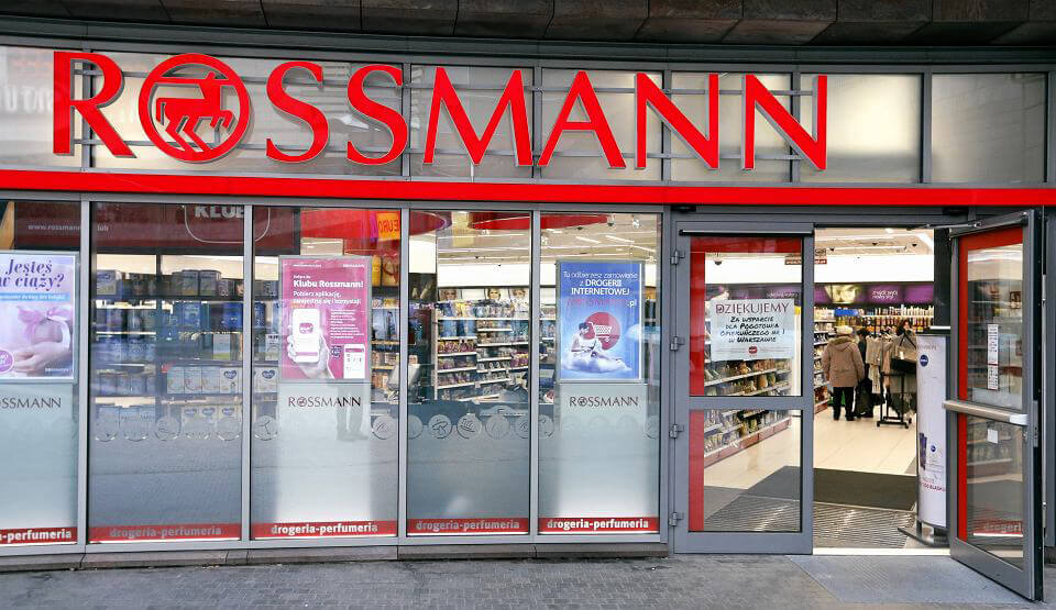 Frontowe wejście do sklepu Rossmann z dużym czerwonym szyldem nad drzwiami. Wewnątrz widoczne są półki z różnymi produktami, a kilku klientów robi zakupy.