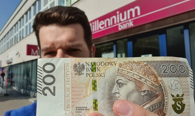 Osoba trzyma przed twarzą banknot o nominale 200 zł przed oddziałem banku Millennium. W tle widoczny jest znak banku.