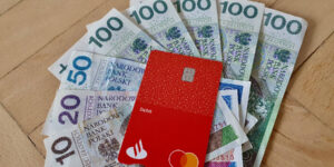 Czerwona karta debetowa jest umieszczona na polskich banknotach o różnych nominałach, w tym 100, 50, 20 i 10 złotych, rozłożonych na drewnianej powierzchni.