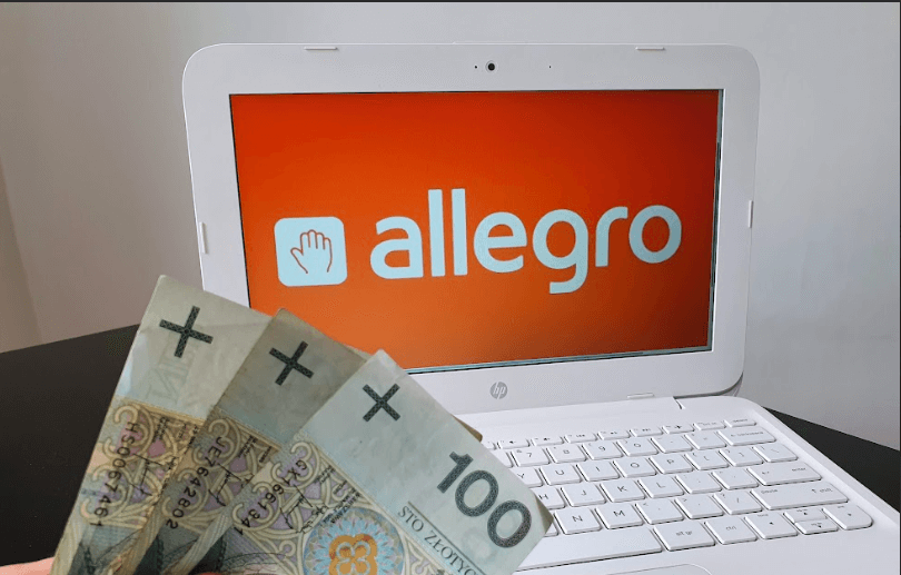 Ręka trzyma trzy banknoty o nominale 100 złotych przed białym laptopem z logo Allegro na pomarańczowym ekranie.