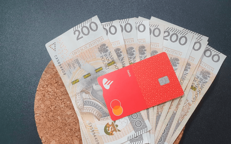 Czerwona karta kredytowa umieszczona jest na kilku banknotach o nominale 200 złotych rozłożonych na korkowej macie, a wszystko to na ciemnym tle.