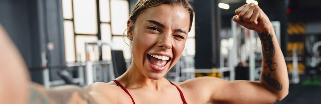 Kobieta w stroju treningowym robi sobie selfie na siłowni, zginając ramię i uśmiechając się entuzjastycznie z jednym okiem zamkniętym. W tle widoczny jest sprzęt do ćwiczeń.