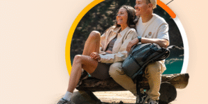 Uśmiechnięta para siedzi razem na pniu na świeżym powietrzu, ubrana swobodnie na wędrówkę. Kobieta obejmuje mężczyznę ramieniem, a obok nich spoczywa plecak. Kolorowy pomarańczowy i żółty łuk zdobi tło.
