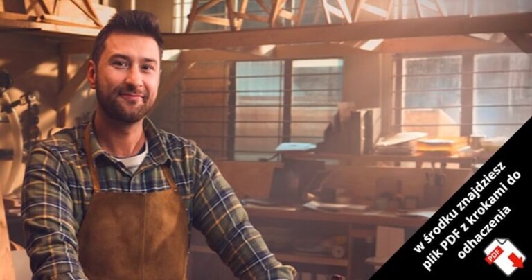 Uśmiechnięty mężczyzna z brodą, ubrany w kraciastą koszulę i brązowy fartuch, stoi w oświetlonym słońcem warsztacie stolarskim. W prawym dolnym rogu znajduje się czarny baner z białym polskim tekstem i czerwoną ikoną PDF.