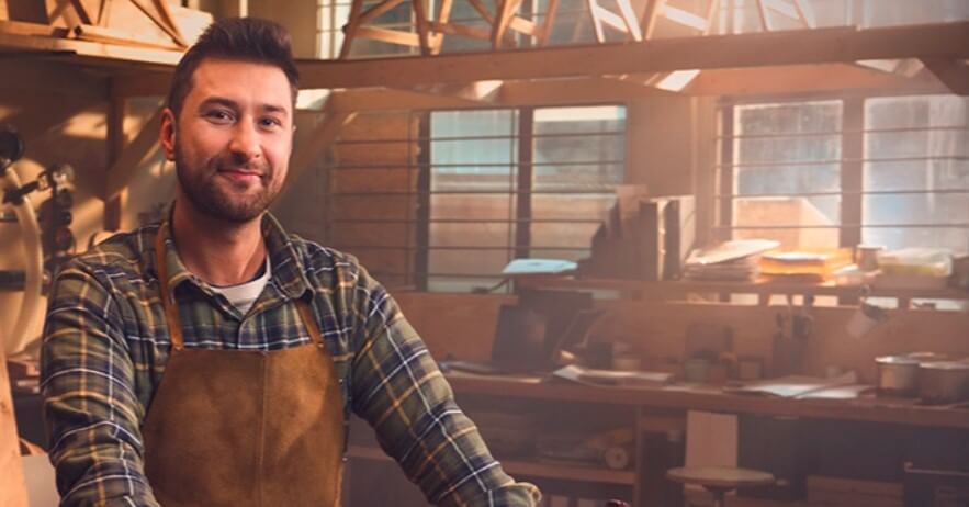 Mężczyzna w koszuli w kratę i brązowym fartuchu uśmiecha się, stojąc w oświetlonym słońcem warsztacie stolarskim wypełnionym narzędziami, drewnianymi półkami i różnymi materiałami.