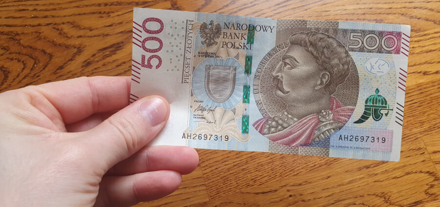 Ręka trzymająca polski banknot o nominale 500 zł, przedstawiający portret mężczyzny z wąsami i w zbroi, na tle drewnianej powierzchni.