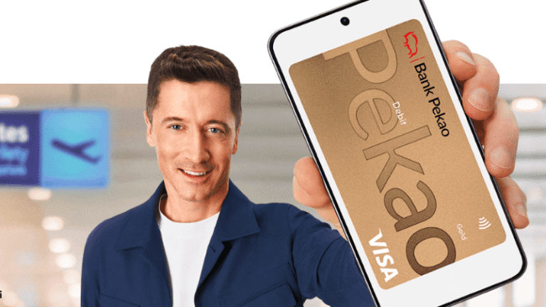 Uśmiechnięty mężczyzna w niebieskiej koszuli trzyma smartfon z cyfrową złotą kartą Visa Banku Pekao. Znak bramy lotniska i rozmyte tło sugerują scenerię podróży.