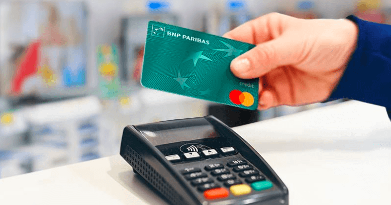 Ręka trzyma kartę kredytową BNP Paribas Mastercard nad terminalem płatniczym, wskazując na płatność zbliżeniową. Tło jest rozmyte, skupiając się na karcie i urządzeniu płatniczym.