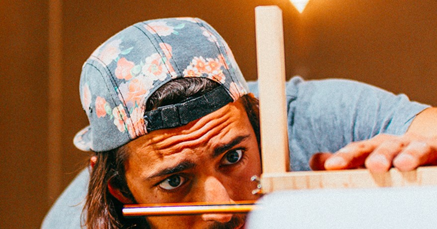 Osoba w kwiecistej czapce bejsbolowej z daszkiem z tyłu uważnie wyrównuje ołówek na drewnianej konstrukcji, koncentrując się na swojej pracy. Ciepłe oświetlenie tworzy skoncentrowaną atmosferę.