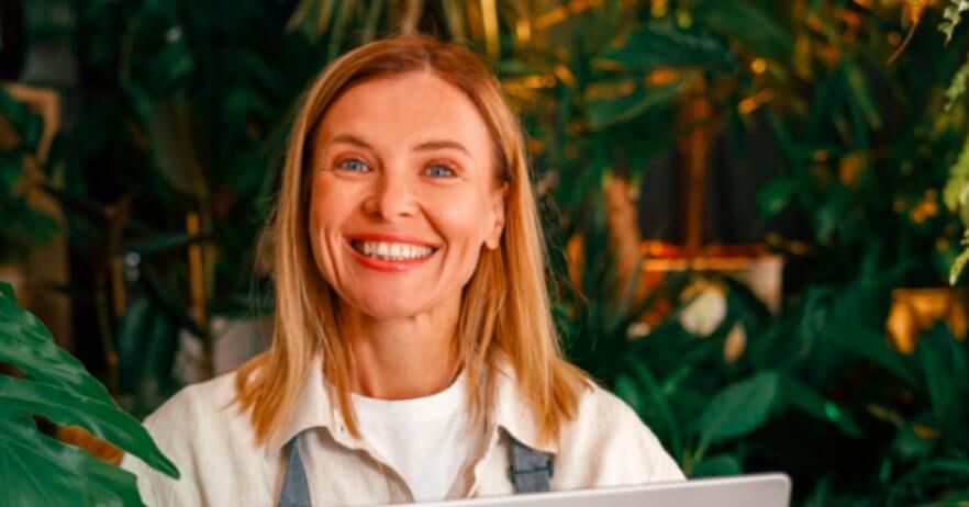 Uśmiechnięta kobieta z blond włosami i białym topem siedzi otoczona zielonymi roślinami, trzymając laptopa. Tło wypełnione jest bujnymi liśćmi, co daje naturalną, radosną atmosferę.