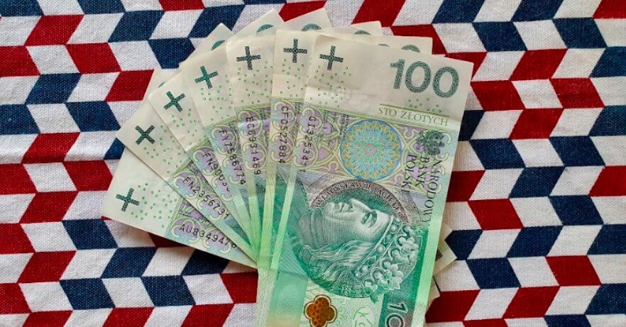 Wachlarz z sześciu banknotów o nominale 100 złotych, umieszczonych na wzorzystej tkaninie o geometrycznych kształtach w kolorach czerwonym, białym i niebieskim.