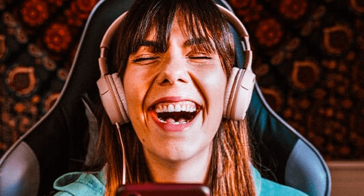 Kobieta z długimi włosami i słuchawkami siedzi na krześle, uśmiechając się szeroko i śmiejąc się, patrząc na telefon. Tło stanowi ciemny, wzorzysty gobelin.