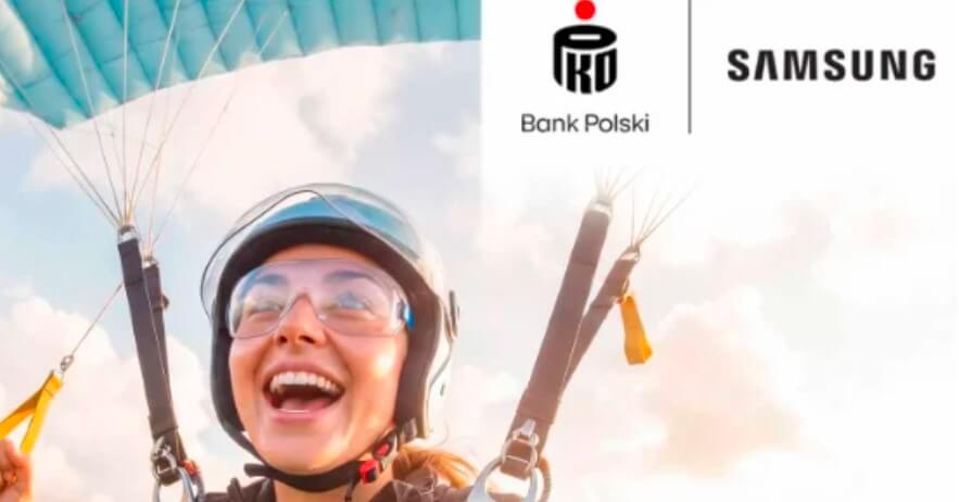 Uśmiechnięta osoba w kasku i goglach skacze ze spadochronem. W prawym górnym rogu, na tle jasnego nieba, widoczne są logo PKO Bank Polski i Samsunga.