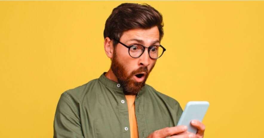 Mężczyzna w okularach i z brodą wygląda na zaskoczonego, trzymając smartfon na żółtym tle.