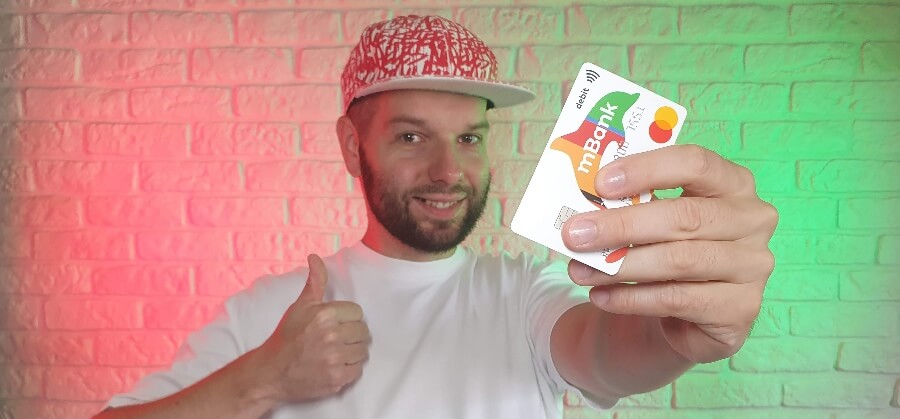 Mężczyzna w czapce w wzory pokazuje kciuk w górę, trzymając przed obiektywem kartę debetową mBanku.